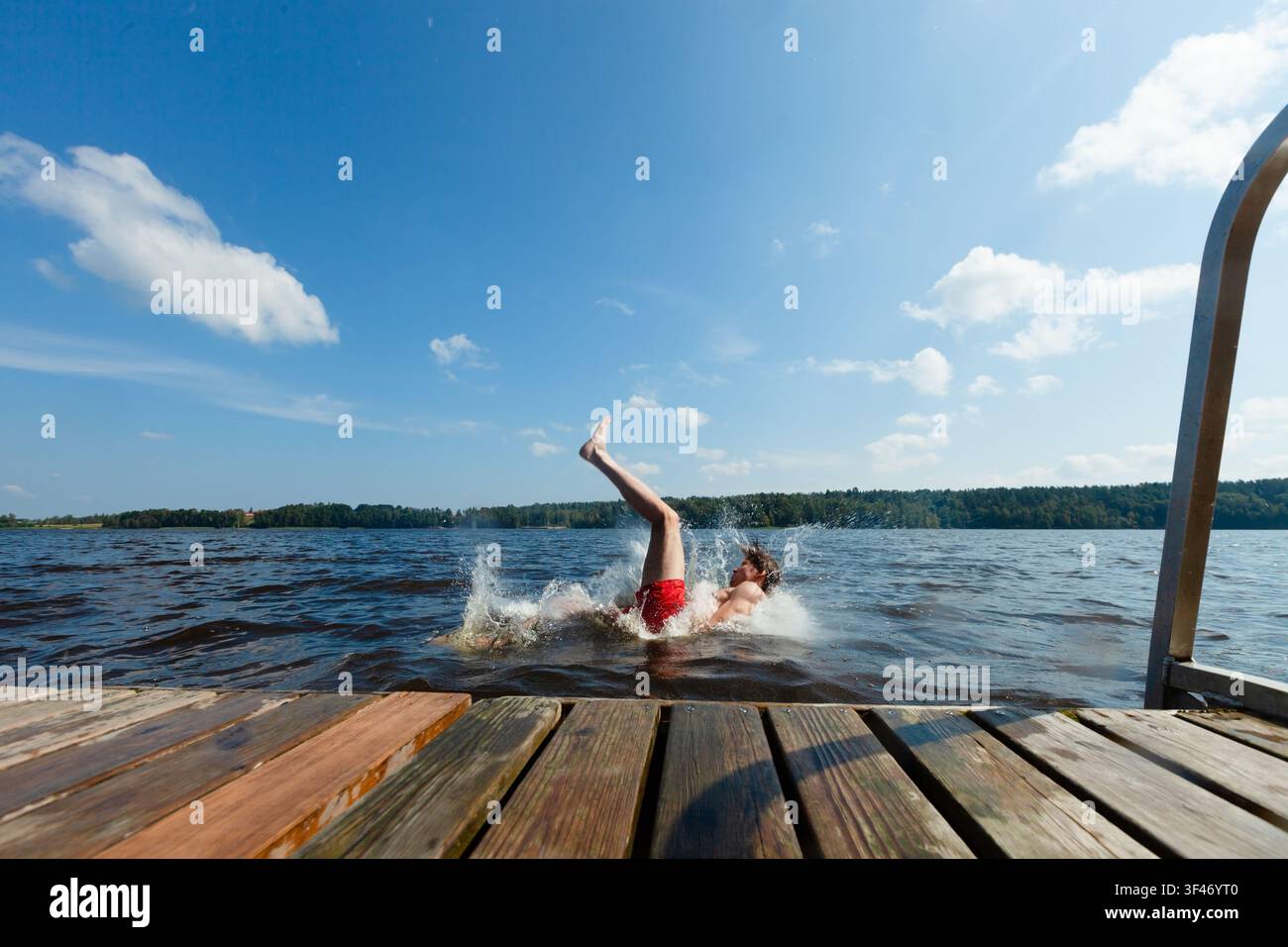 Un homme tombe d'une jetée en bois dans un lac en éclaboussant de l'eau par une journée d'été. Un gars perd l'équilibre et culbute du bord dans l'eau. Un adulte vit un imprévu en plein air et un accident comique au lac Banque D'Images