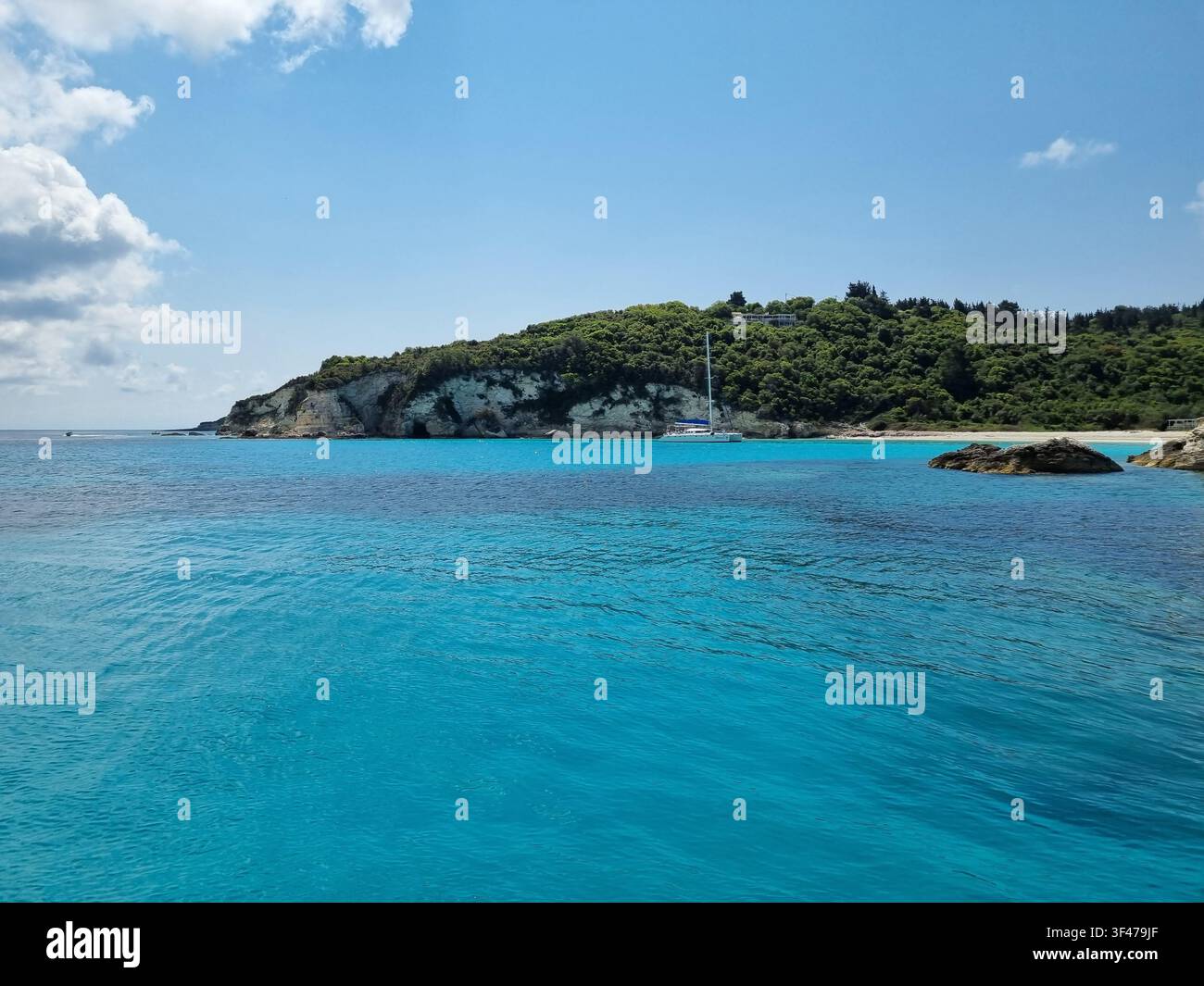 Catamaran ancré devant une plage de galets blancs dans une baie turquoise avec des falaises calcaires boisées, Antipaxos, Îles Ioniennes, Grèce - Image de stock capturée avec un smartphone
