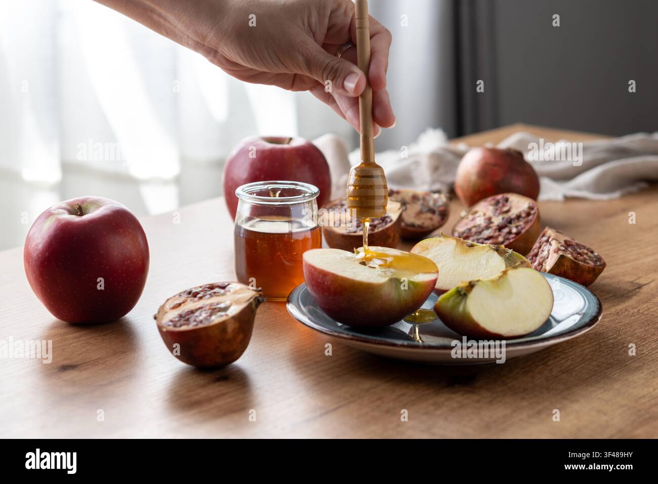Main plongeant une tranche de pomme dans du miel à côté d’un grenade, d’un pot et de pommes entières sur une table en bois sous une lumière douce, symbolisant Rosh Hashanah, le Nouvel An juif Banque D'Images