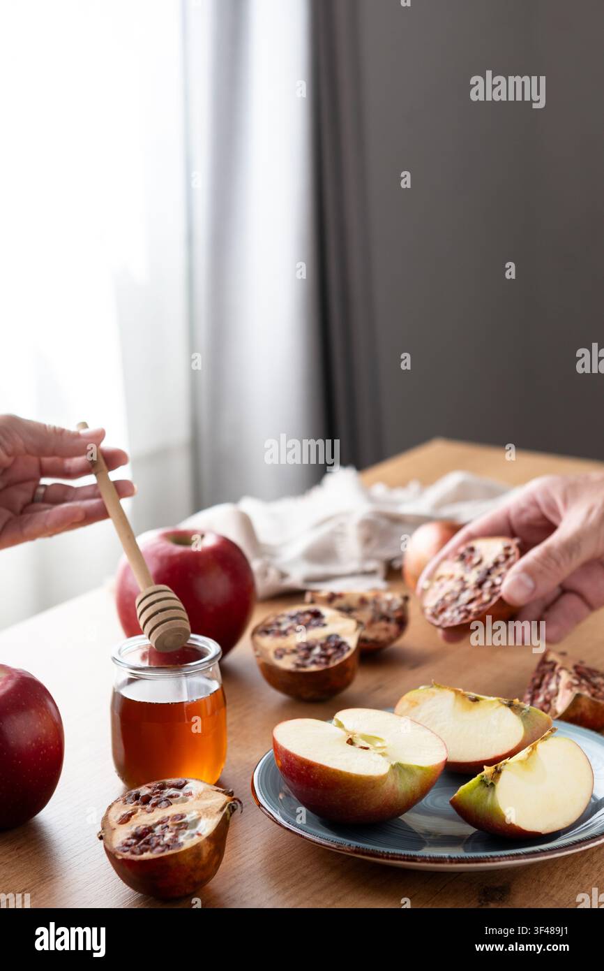 Close-up de mains préparant la célébration de Rosh Hashanah avec des tranches de pomme, un cuillère à miel et un pot, ainsi que des moitiés de grenade sur une table, symbolisant une douceur Banque D'Images