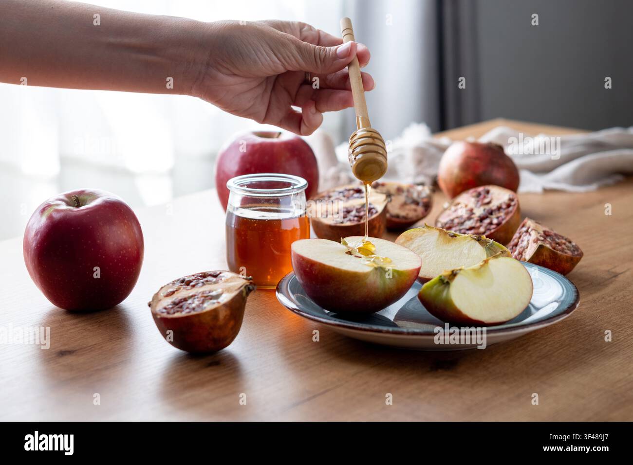 Main plongeant des tranches de pomme dans du miel à côté d'un pot, pommes fraîches et grenade coupée sur une table, célébration traditionnelle juive du Nouvel An Rosh Hashanah Banque D'Images