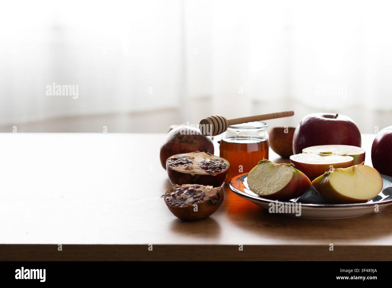 Nature morte minimaliste de pommes, pot de miel avec cuillère et grenade sur une assiette dans une lumière douce de fenêtre, symbolisant Rosh Hashanah et le Nouvel An juif Banque D'Images