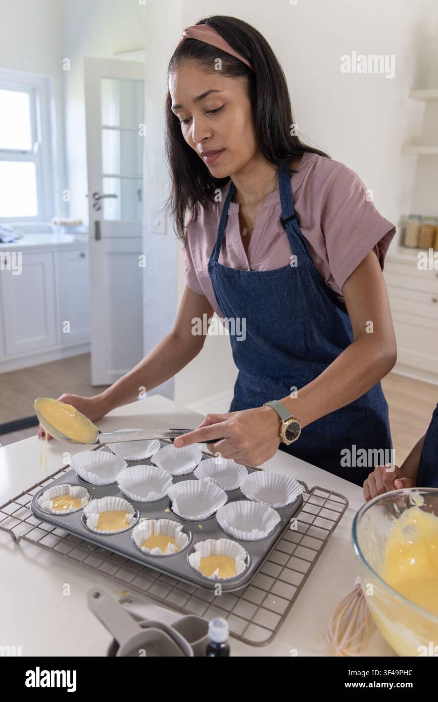 Mère afro-américaine en tablier en jean versant la pâte dans un moule à muffins tandis qu'un jeune tient un bol dans la cuisine Banque D'Images