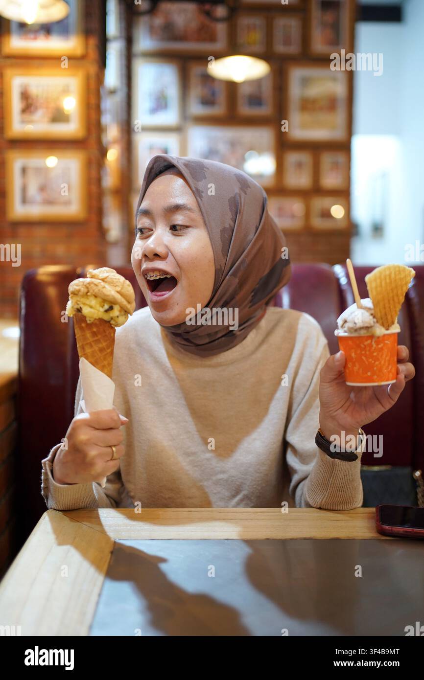 Femme indonésienne en hijab tenant une glace en cornet et une tasse tout en savourant un dessert dans un café. Concept de nourriture amusante et de style de vie. Banque D'Images