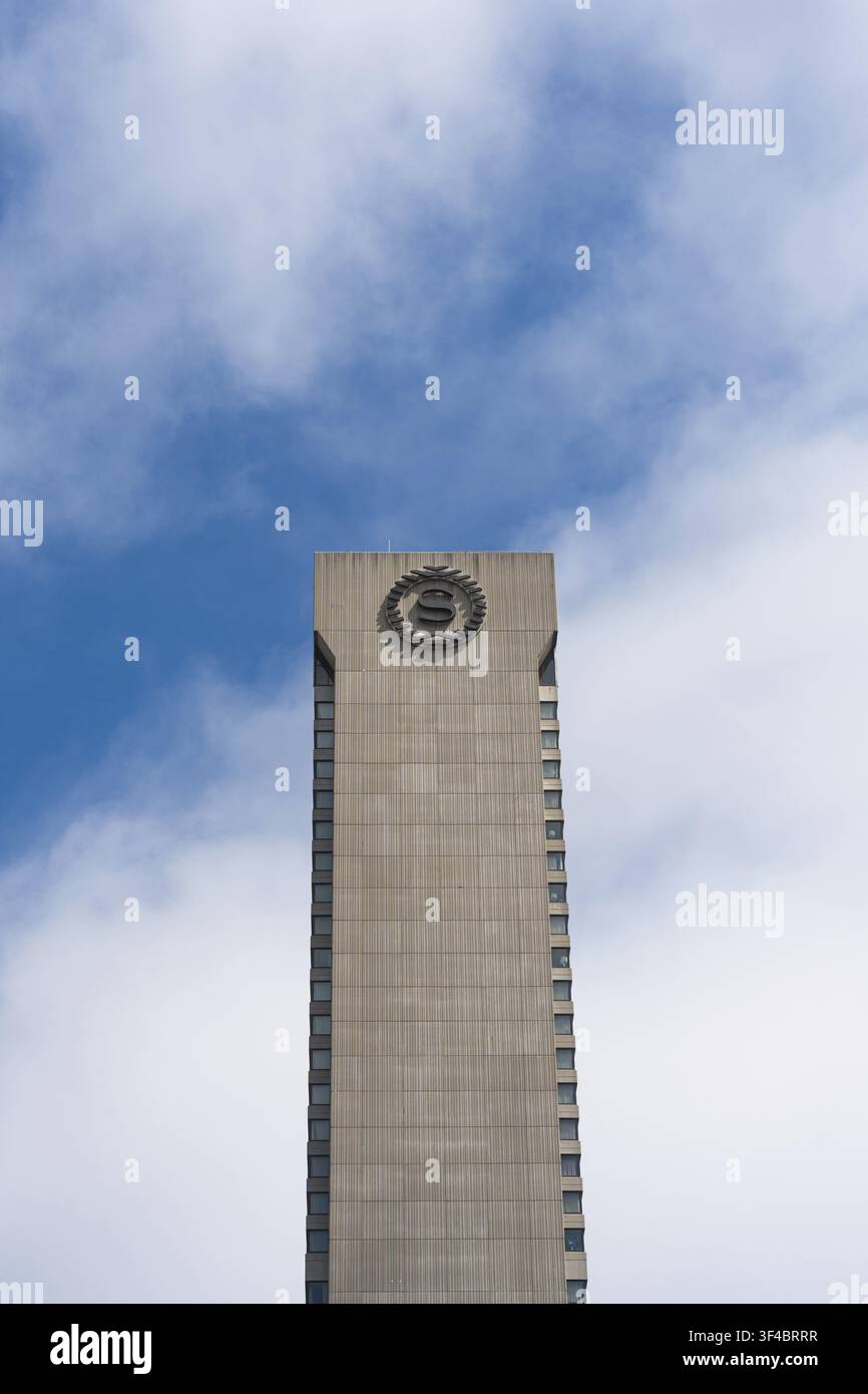 Horloge tournante contre un ciel bleu nuageux, façade en béton austère s'élevant hors du cadre, perspective verticale mettant l'accent sur le modernisme minimaliste Banque D'Images