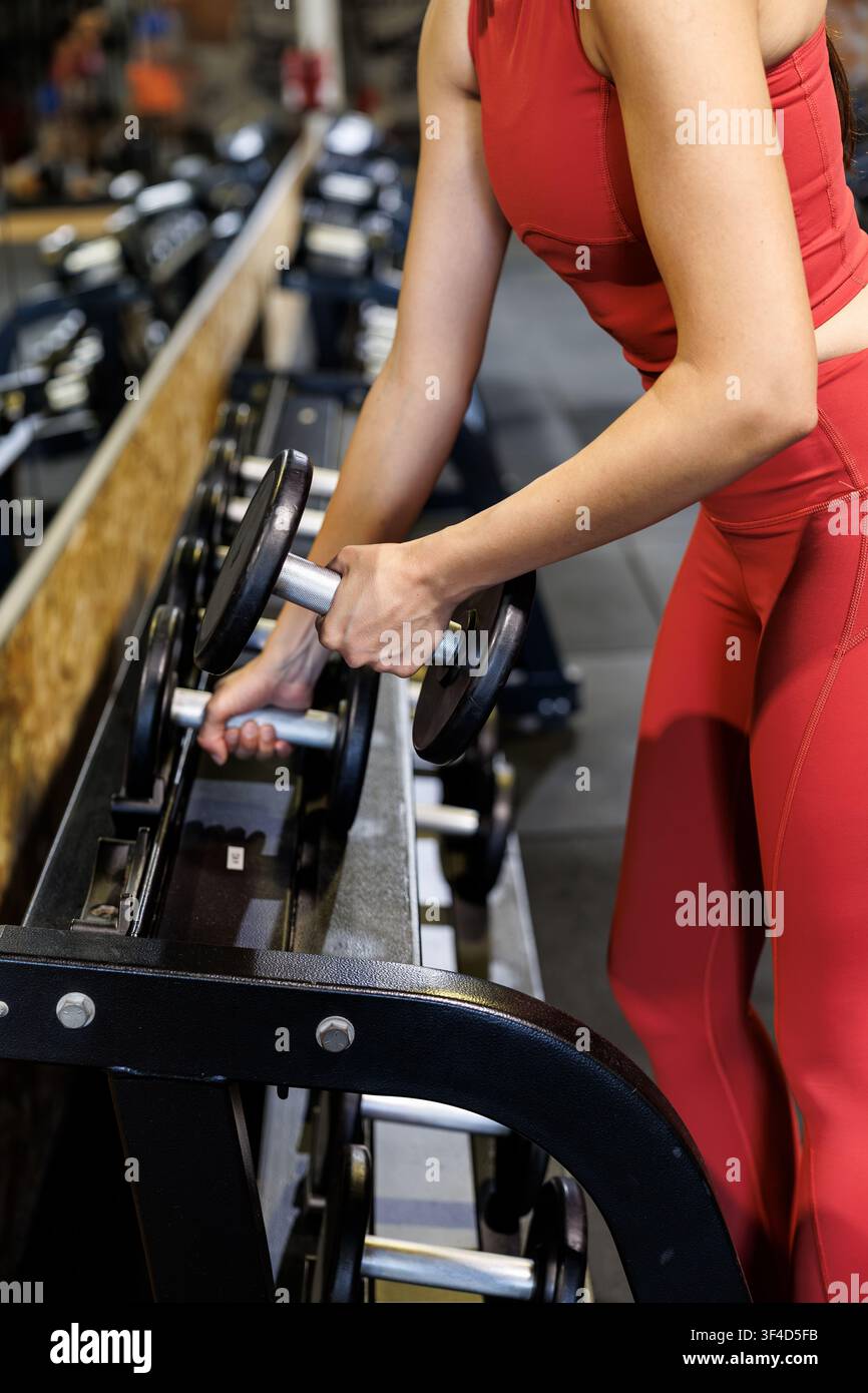 Une femme soulève des poids dans un cadre de salle de sport. Elle est concentrée sur sa routine d'entraînement. La salle de sport est équipée de divers appareils d'exercice. Banque D'Images