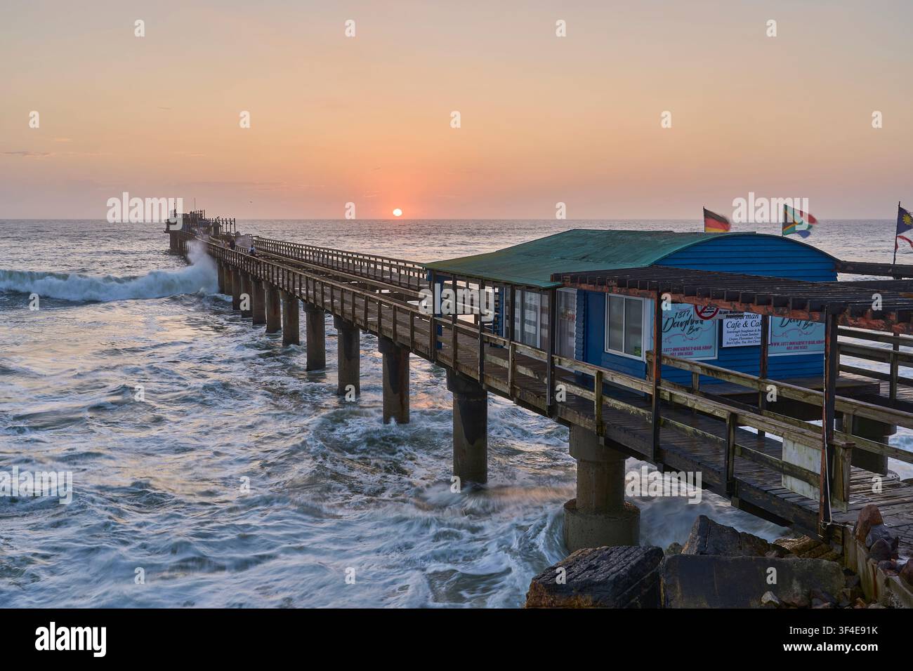 Un coucher de soleil doré éclatant encadre la jetée en bois historique de Swakopmund, en Namibie, alors que les puissantes vagues de l'océan Atlantique s'écrasent contre ses piliers. Banque D'Images
