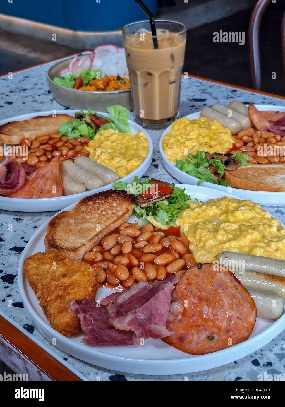 Close-up d'une assiette de petit-déjeuner anglais traditionnel comprenant des œufs brouillés, des haricots cuits au four, des saucisses, du pain grillé et une hash brown sur une table en terrazzo. Banque D'Images