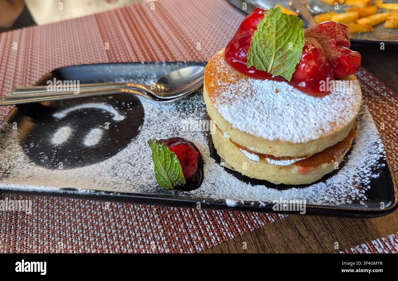 Dessert de pancake souflé japonais garni de sucre en poudre, de cerises et de feuilles de menthe, servi dans une assiette en céramique noire, dans un café. Banque D'Images