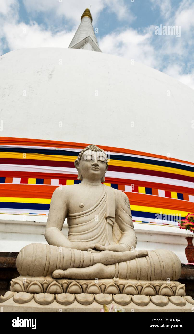 Statue de Bouddha en pierre assise devant une grande stûpa bouddhiste blanche enveloppée dans une bannière de drapeau bouddhiste colorée, Sri Lanka. Banque D'Images