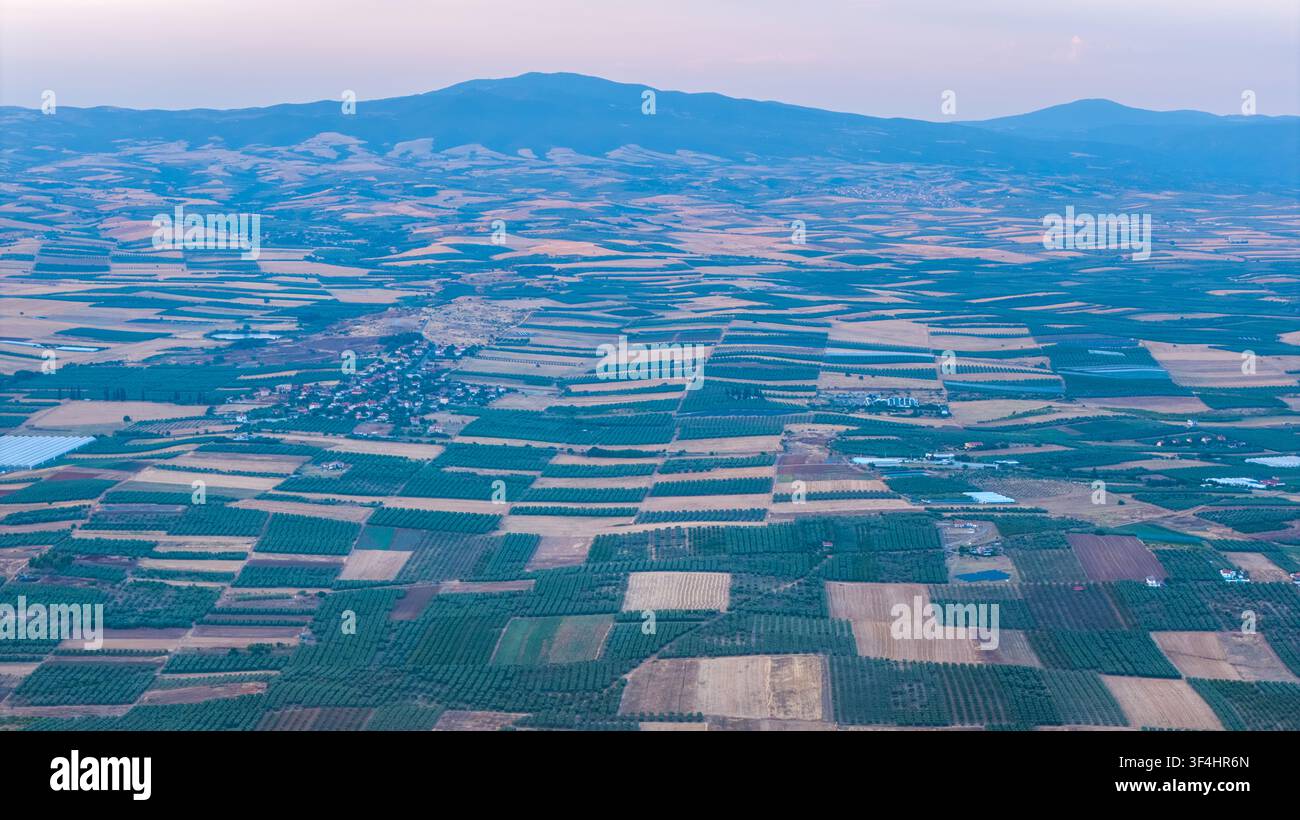 Une vue panoramique élevée montre des champs cultivés en blocs géométriques, une petite colonie et des crêtes montagneuses superposées sous un ciel pastel doux du soir. Banque D'Images