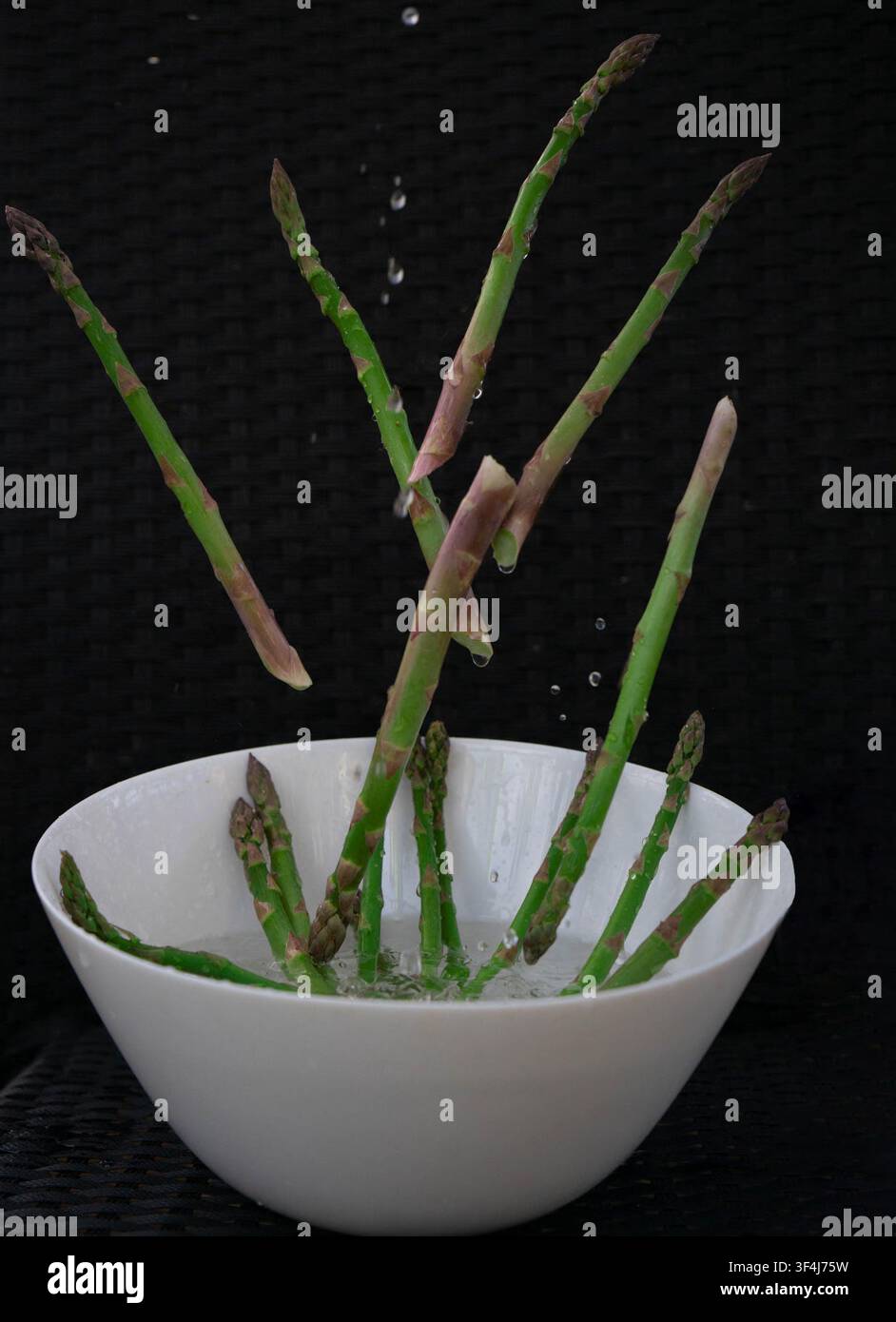 Des pointes d'asperges vertes fraîches tombant dans un bol en céramique blanc sur fond noir, capturées en mouvement comme un concept d'alimentation saine de saison. Banque D'Images