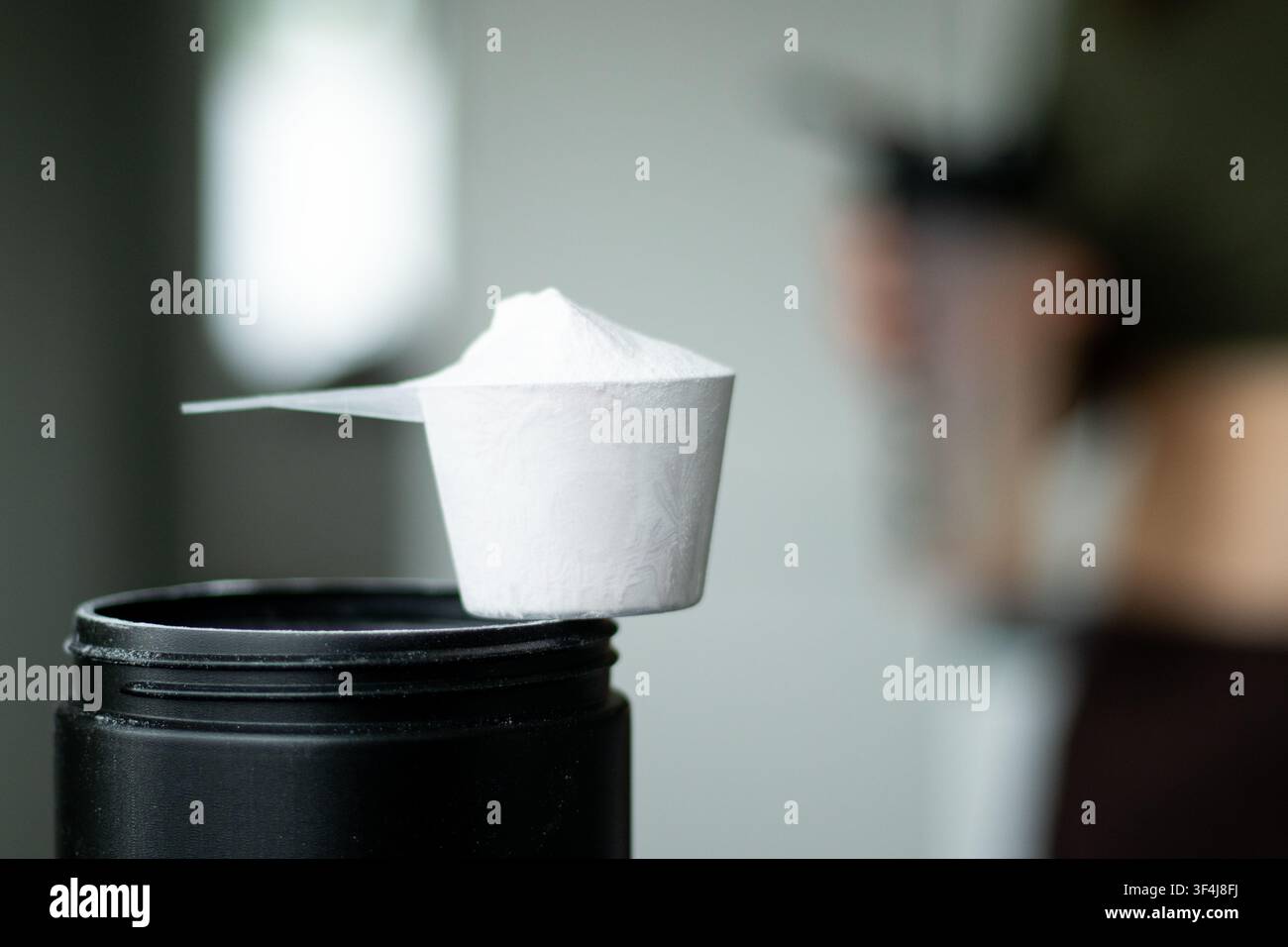 Cuillère à poudre protéinée blanche reposant sur un récipient noir avec un fond flou d'une personne tenant une bouteille shaker dans un environnement de fitness Banque D'Images