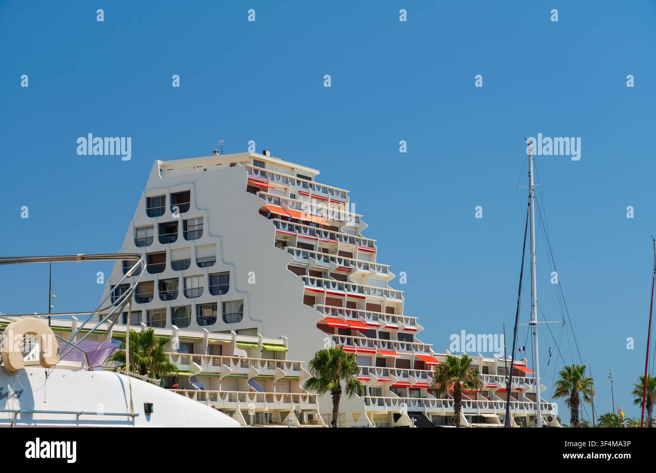 Bâtiment pyramide blanc emblématique à La Grande-Motte avec des auvents rouges sous un ciel bleu clair, vu depuis une marina avec des palmiers Banque D'Images