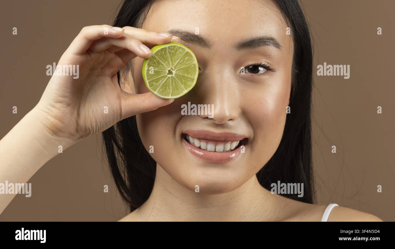 Femme asiatique souriante tenant une tranche de citron vert devant l'œil, gros plan, concept de mode de vie sain Banque D'Images