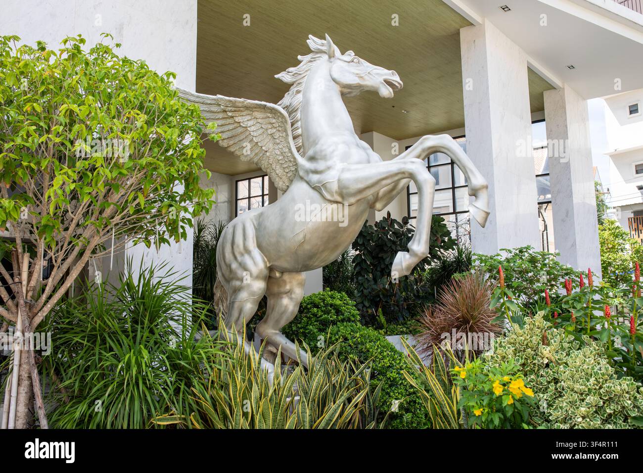 Statue de pégase blanc cabré avec les ailes déployées devant l'entrée du bâtiment entourée de plantes vertes Banque D'Images
