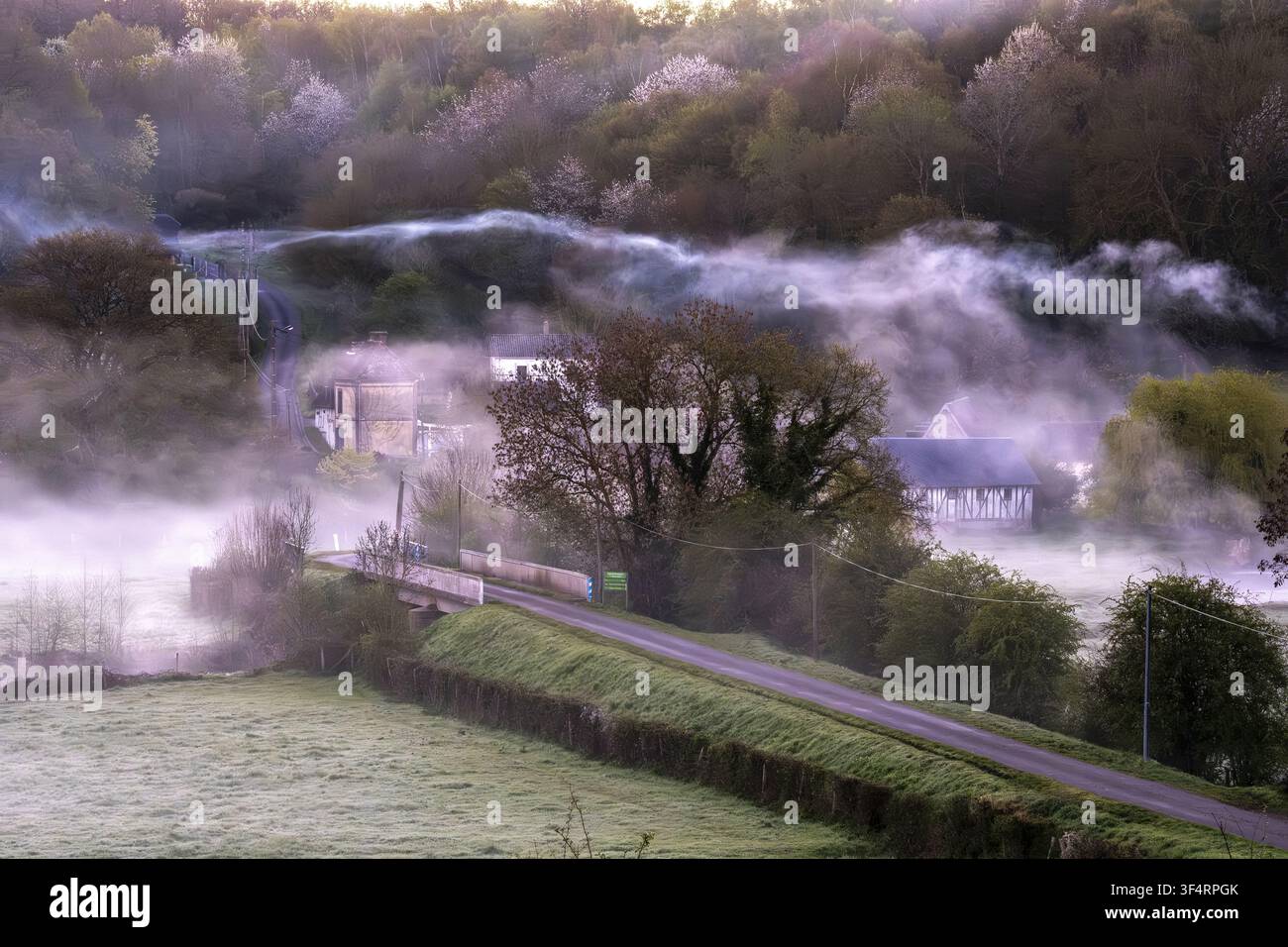 Champignolles, Eure, France : un paysage rural brumeux le matin, avec des maisons, une route et un pont. Banque D'Images