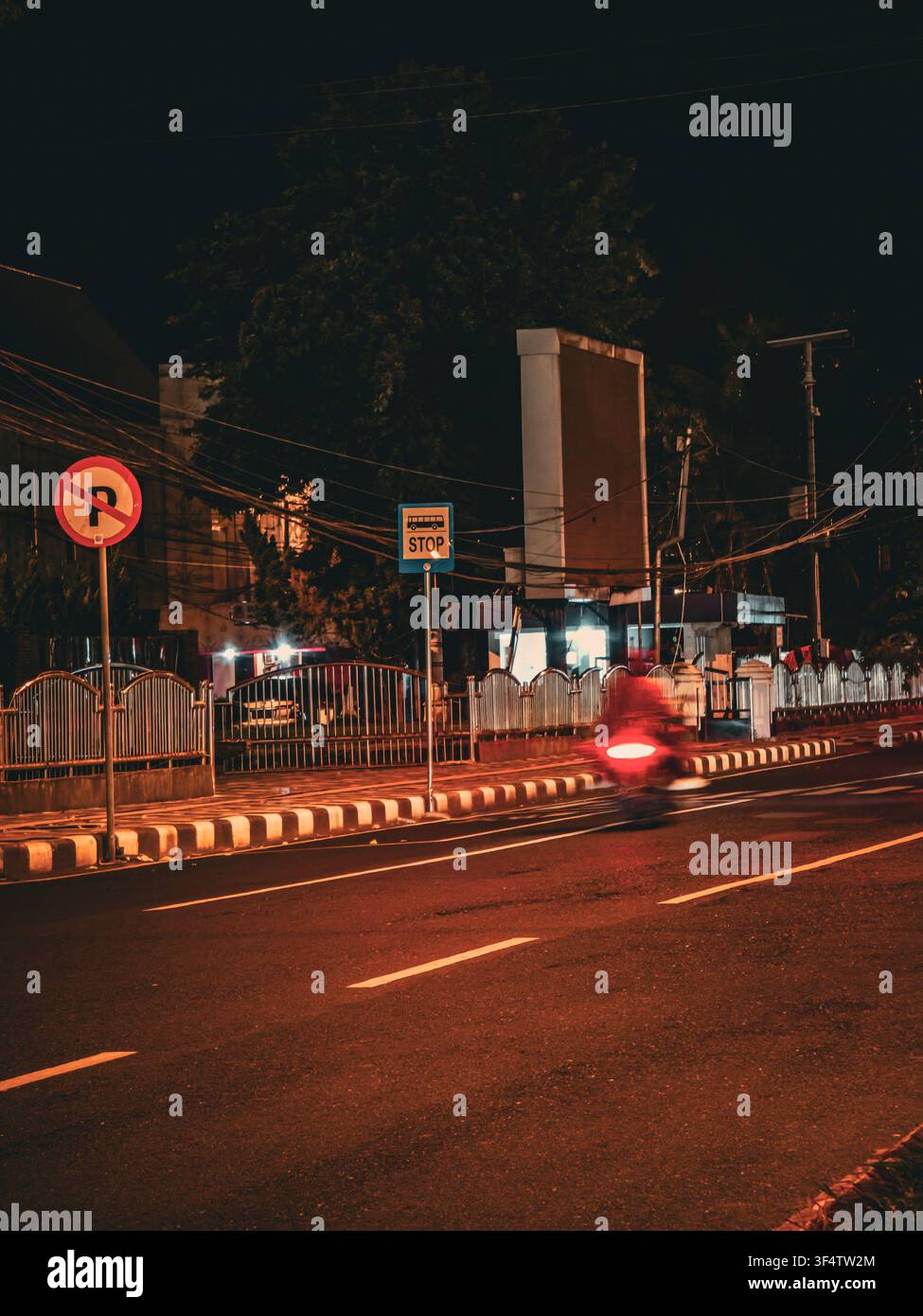 Rue urbaine déserte la nuit avec des panneaux de signalisation, une barrière de bord de route et le flou de mouvement du feu arrière d’une moto qui passe. - Image de stock capturée avec un smartphone