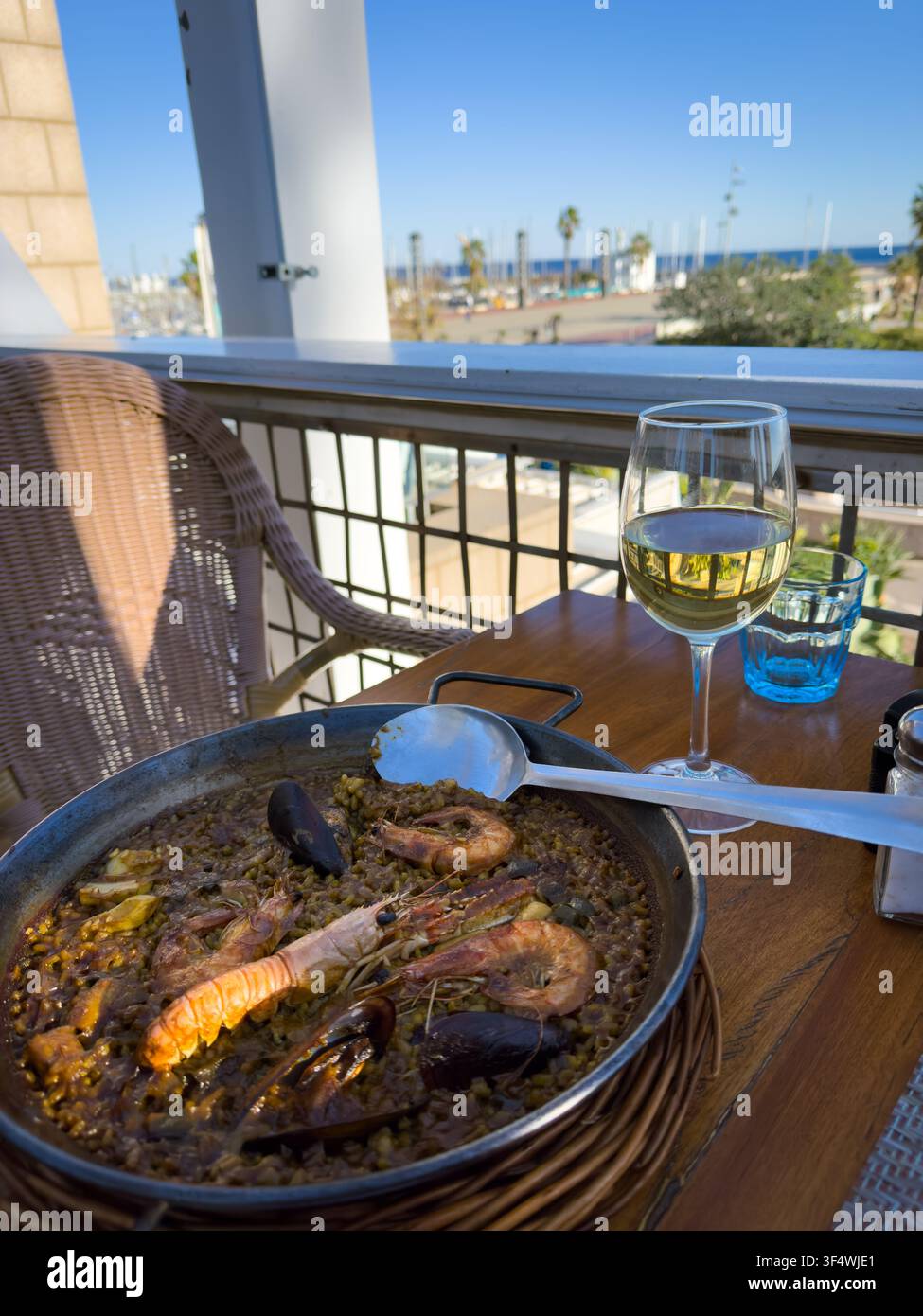 Une paella aux fruits de mer et un verre de vin blanc posent sur une table de restaurant surplombant une marina ensoleillée. Banque D'Images