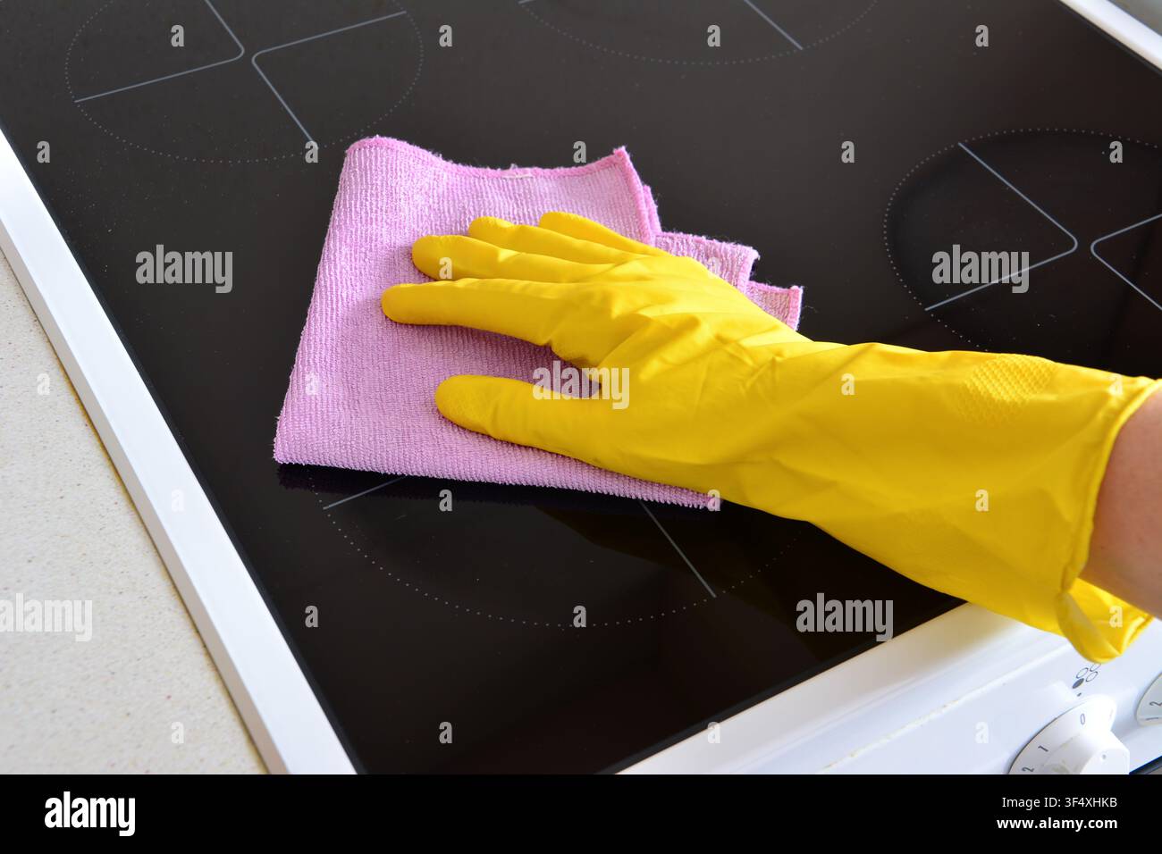 Personne portant un gant jaune nettoyant une plaque de cuisson en céramique noire avec un chiffon rose Banque D'Images
