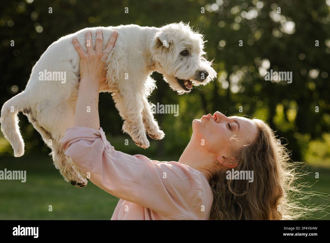 Une femme joyeuse soulève son chien heureux dans les airs, mettant en valeur leur lien affectueux dans un magnifique cadre de parc baigné de lumière. Banque D'Images