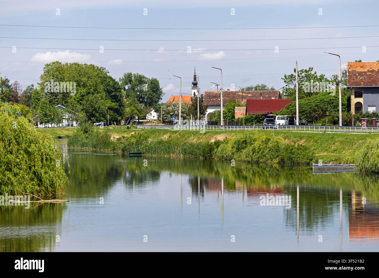 Une scène paisible de village rural avec une rivière calme reflétant des arbres, des bateaux et un clocher d'église à côté d'une route bordée de maisons et de verdure sur un brig Banque D'Images