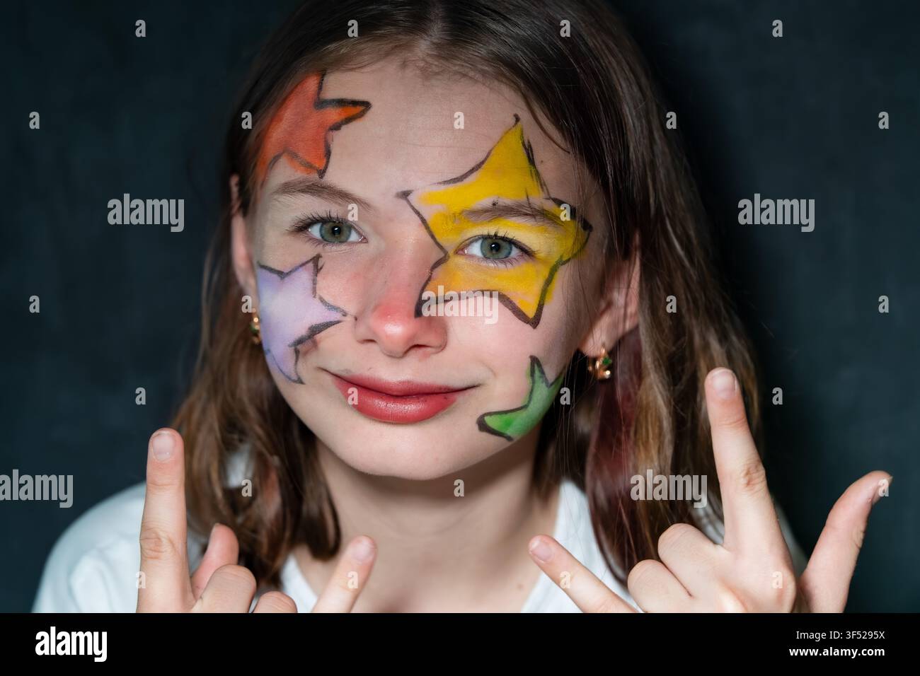 Portrait rapproché d'une préadolescente avec un maquillage facial en forme d'étoiles colorées regardant vers le haut sur un fond sombre Banque D'Images