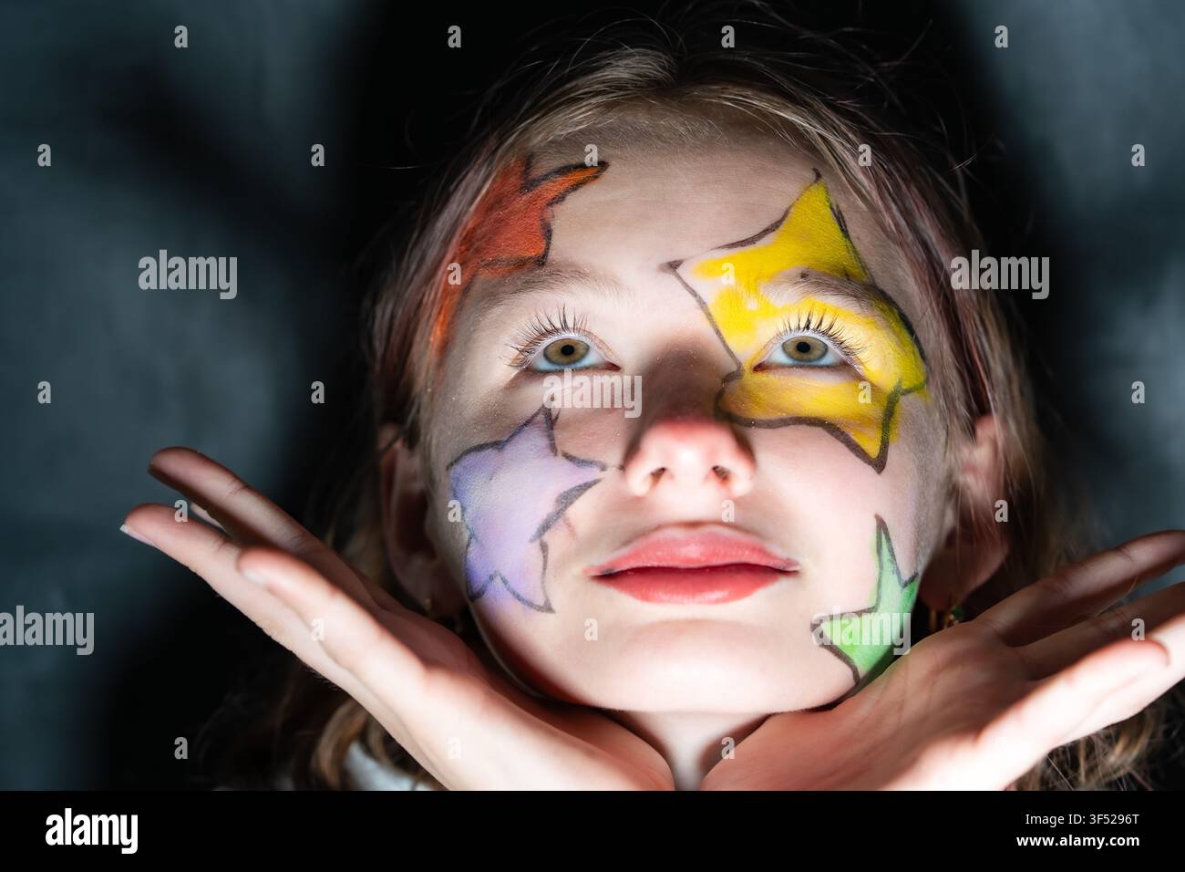 Portrait en gros plan d'une préadolescente avec un maquillage facial en forme d'étoiles colorées, regardant vers le haut et posant les mains sous le menton contre un fond sombre. Banque D'Images
