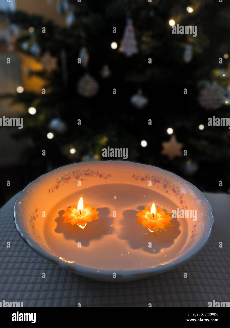 Deux bougies flottantes allumées en forme de fleurs dans un bol d'eau, avec un sapin de Noël flou et des lumières bokeh en arrière-plan. - Image de stock capturée avec un smartphone