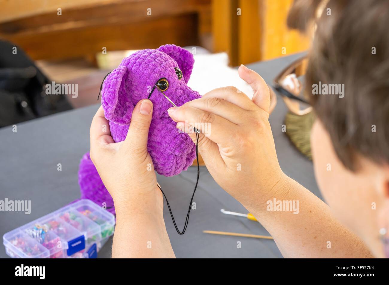 Mains féminines cousant un détail du visage avec une aiguille et du fil noir sur un jouet amigurumi en crochet violet et peluche Banque D'Images