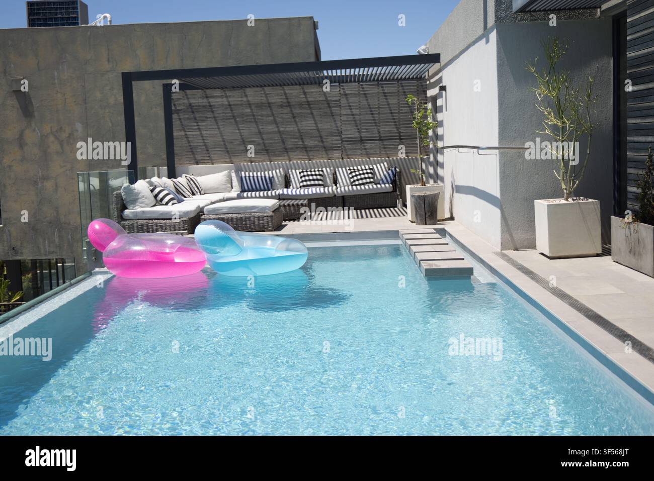 Piscine sur le toit centrée dans le cadre, transats roses et bleus flottant près de la gauche, canapé tissé sous pergola Banque D'Images