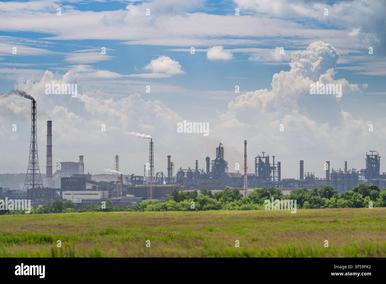 Une vaste usine métallurgique s'étend à l'horizon, marquée par des cheminées imposantes et des structures industrielles. Le ciel est animé de nuages, mettant en valeur Banque D'Images