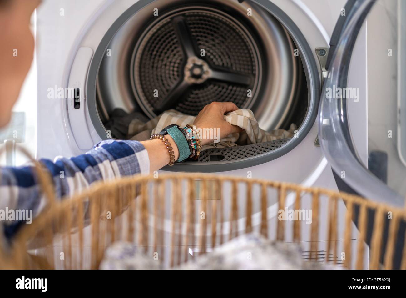 Une femme place un tissu rayé d'un panier en osier dans un sèche-linge à chargement frontal blanc. Une tâche domestique quotidienne. Banque D'Images