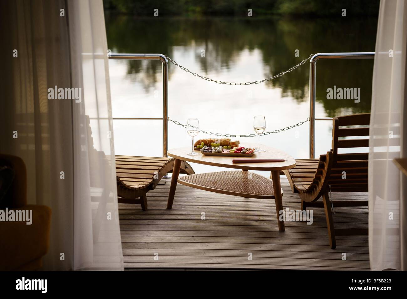 Terrasse en bois d'une péniche avec des snacks et du vin, surplombant une vue calme sur la rivière lors d'un après-midi d'été paisible. Vacances de luxe en glamping, escapade tranquille pour le week-end, dîner en plein air au bord de la rivière Banque D'Images