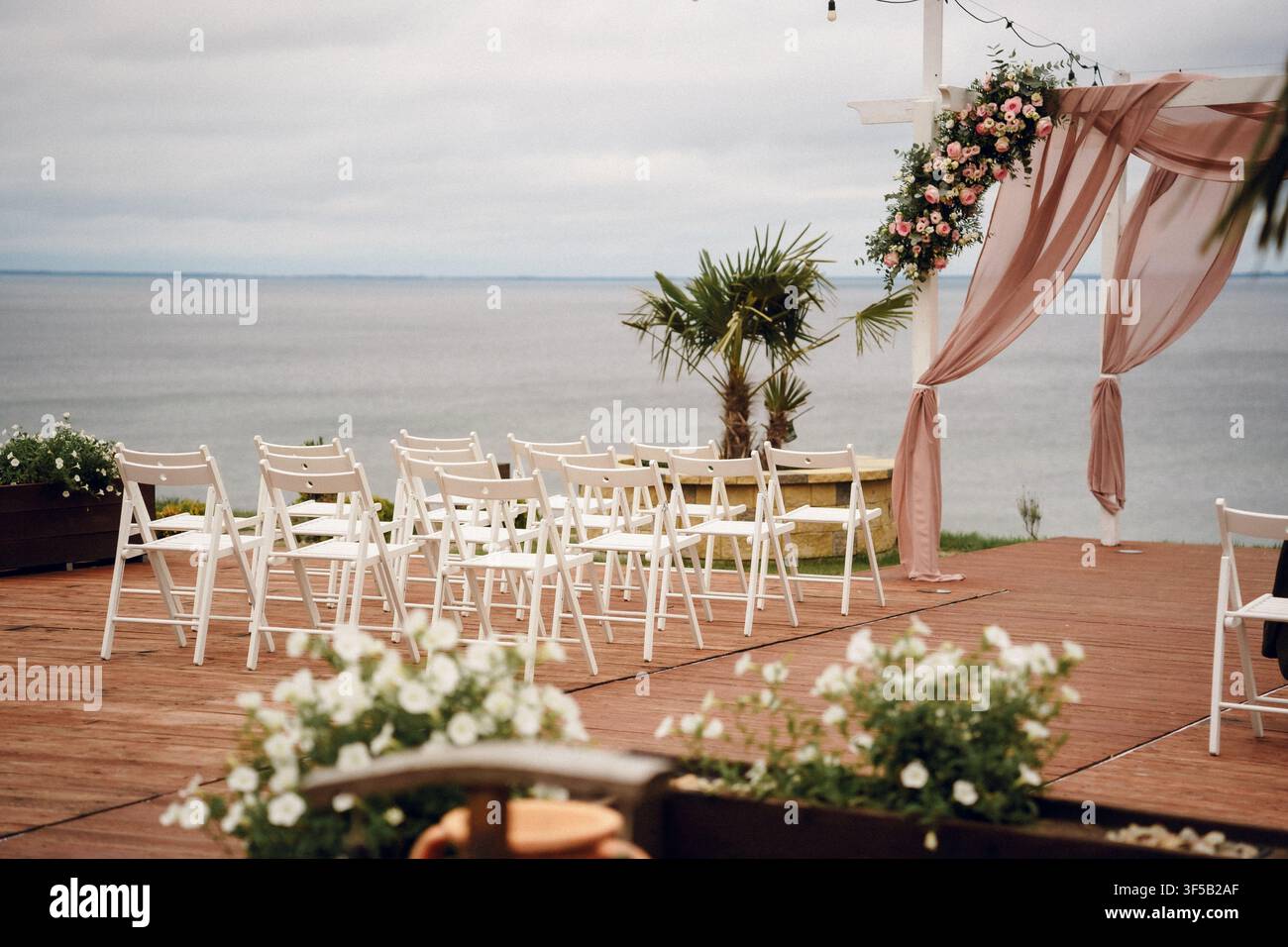 Installation pour une cérémonie de mariage sur la plage en plein air avec des chaises blanches et une arche fleurie surplombant l'océan. Lieu de mariage romantique sur la côte, mariage de destination tropicale, décoration élégante d'autel au bord de la mer Banque D'Images