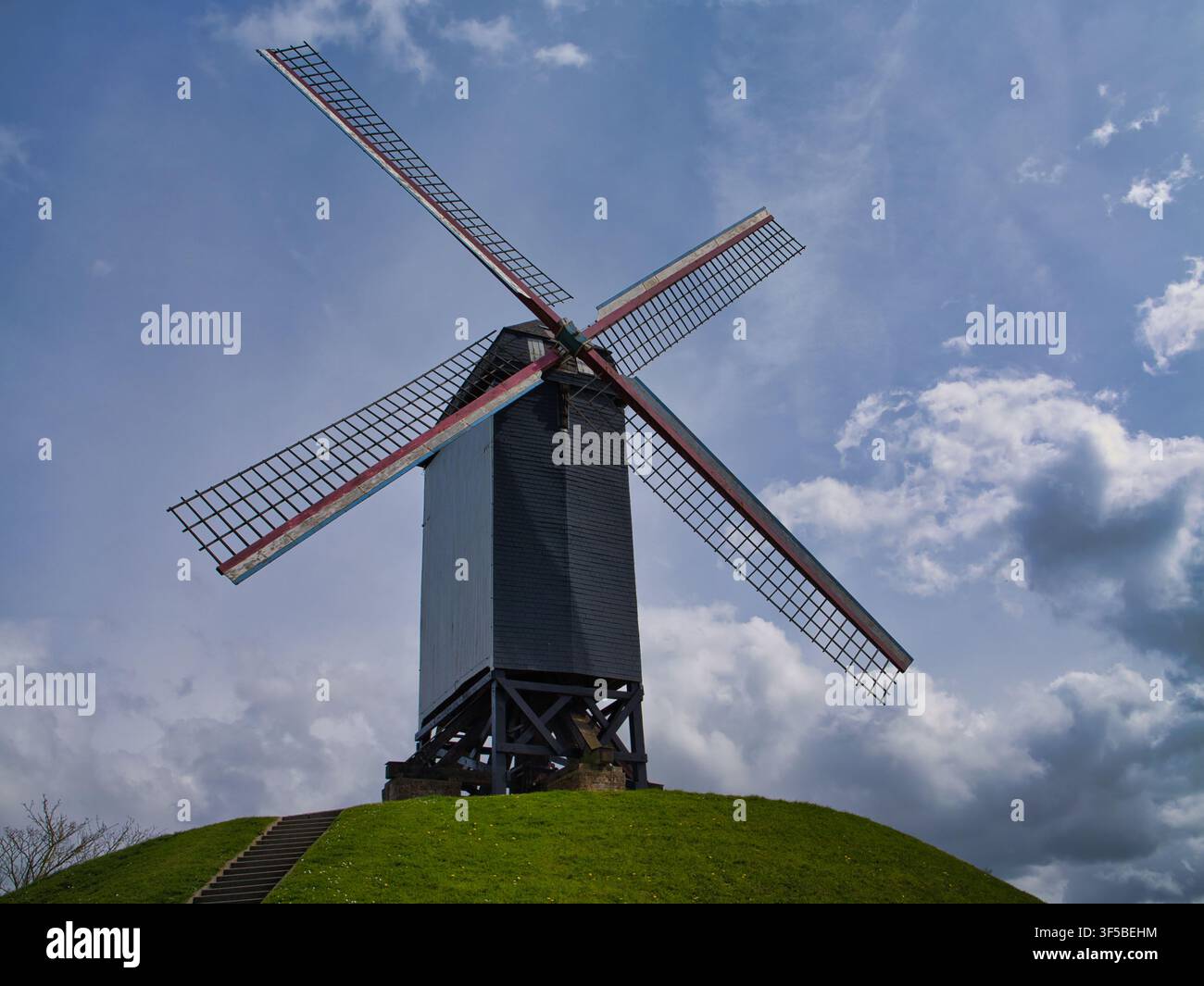 Brugge, Belgique – 10 avr. 2026 : Un moulin à vent traditionnel s’élève au-dessus d’une colline herbeuse à Brugge, en Belgique, sous des nuages dramatiques et un ciel bleu de printemps Banque D'Images