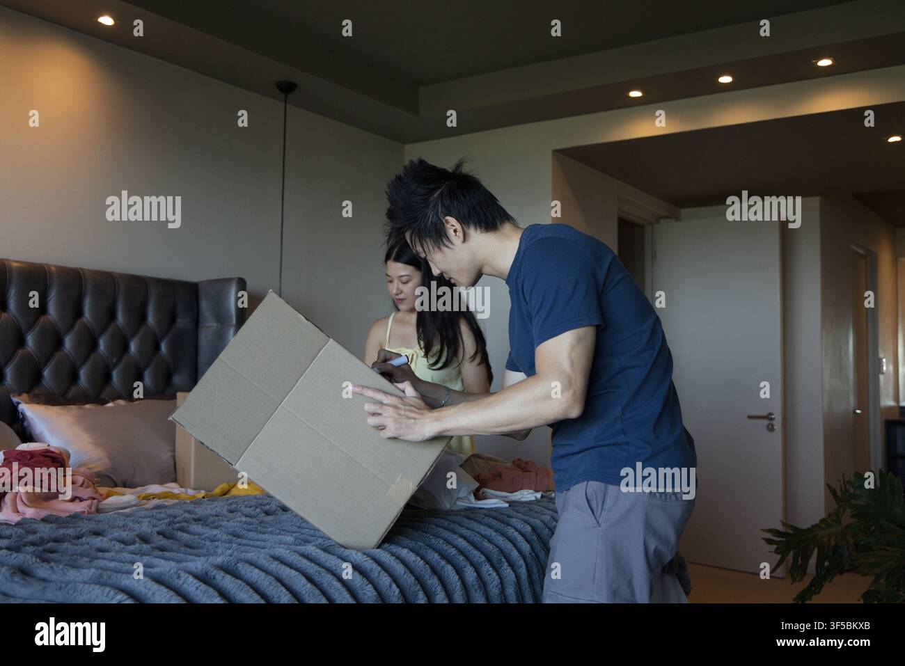 Couple asiatique déballant sur le lit dans la chambre, homme en chemise bleu marine manipulant une boîte, femme vérifiant son smartphone Banque D'Images
