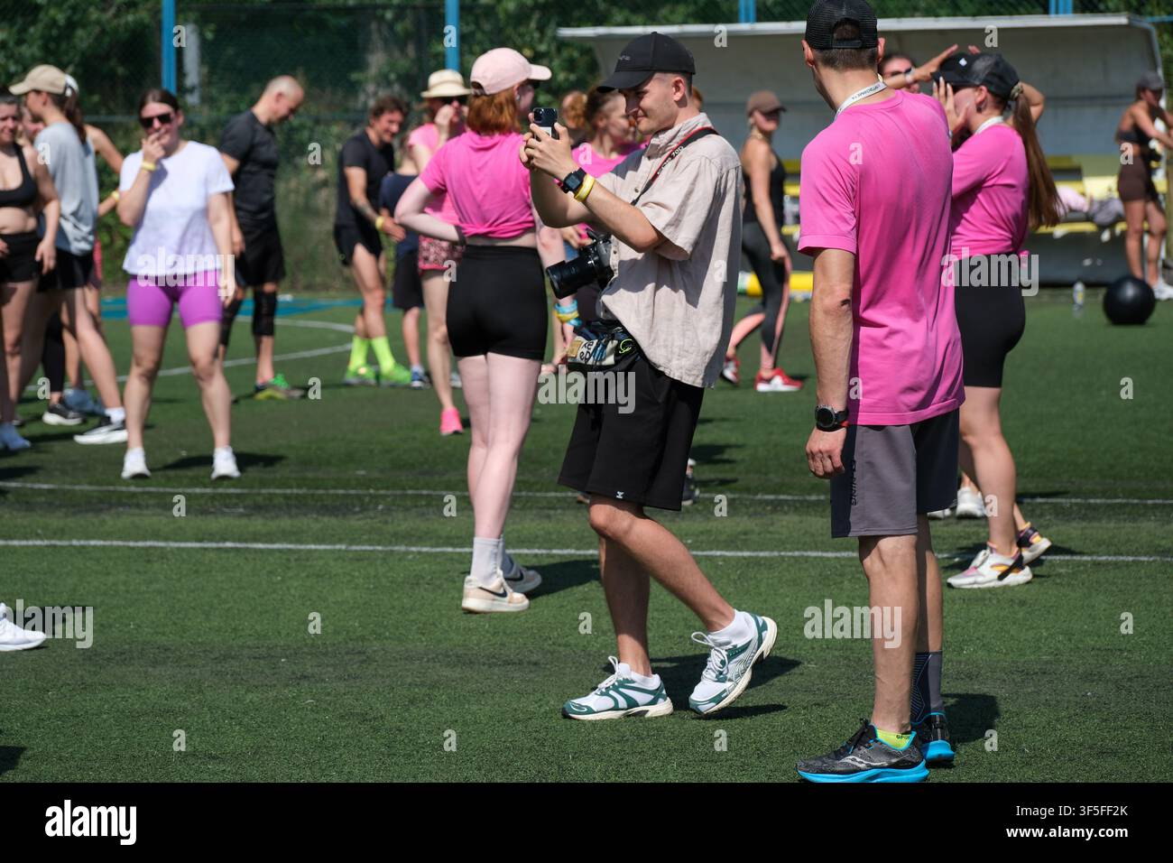 KIEV, UKRAINE - 1ER AOÛT 2026 : Photographe enregistre les participants lors d'un événement de fitness de groupe en plein air sur un terrain de football en gazon synthétique alors que des gens Banque D'Images