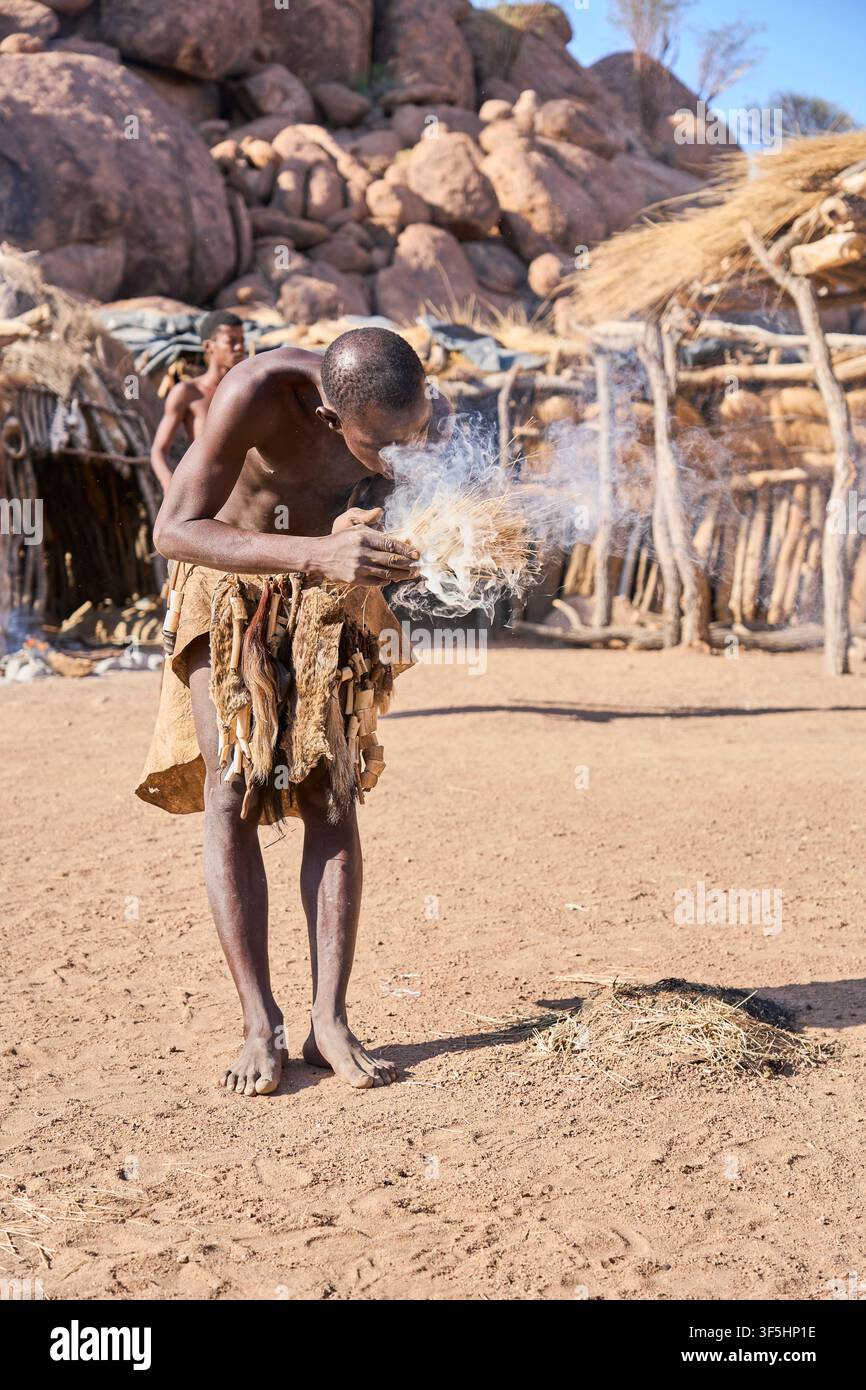 Un homme tribal africain portant des vêtements traditionnels en pagne se tient devant des huttes en paille, insufflant la vie dans un nid d'herbe fumant pour allumer un feu. Banque D'Images