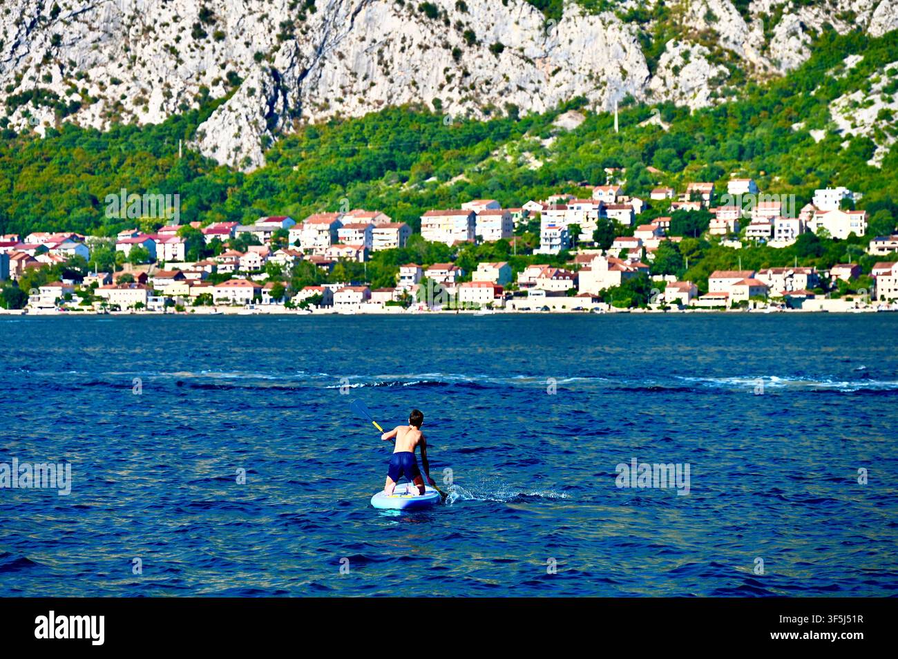 Une personne faisant du paddle sur une mer d'un bleu éclatant, avec un charmant village côtier et d'immenses montagnes rocheuses couvertes de végétation verte en arrière-plan. Banque D'Images