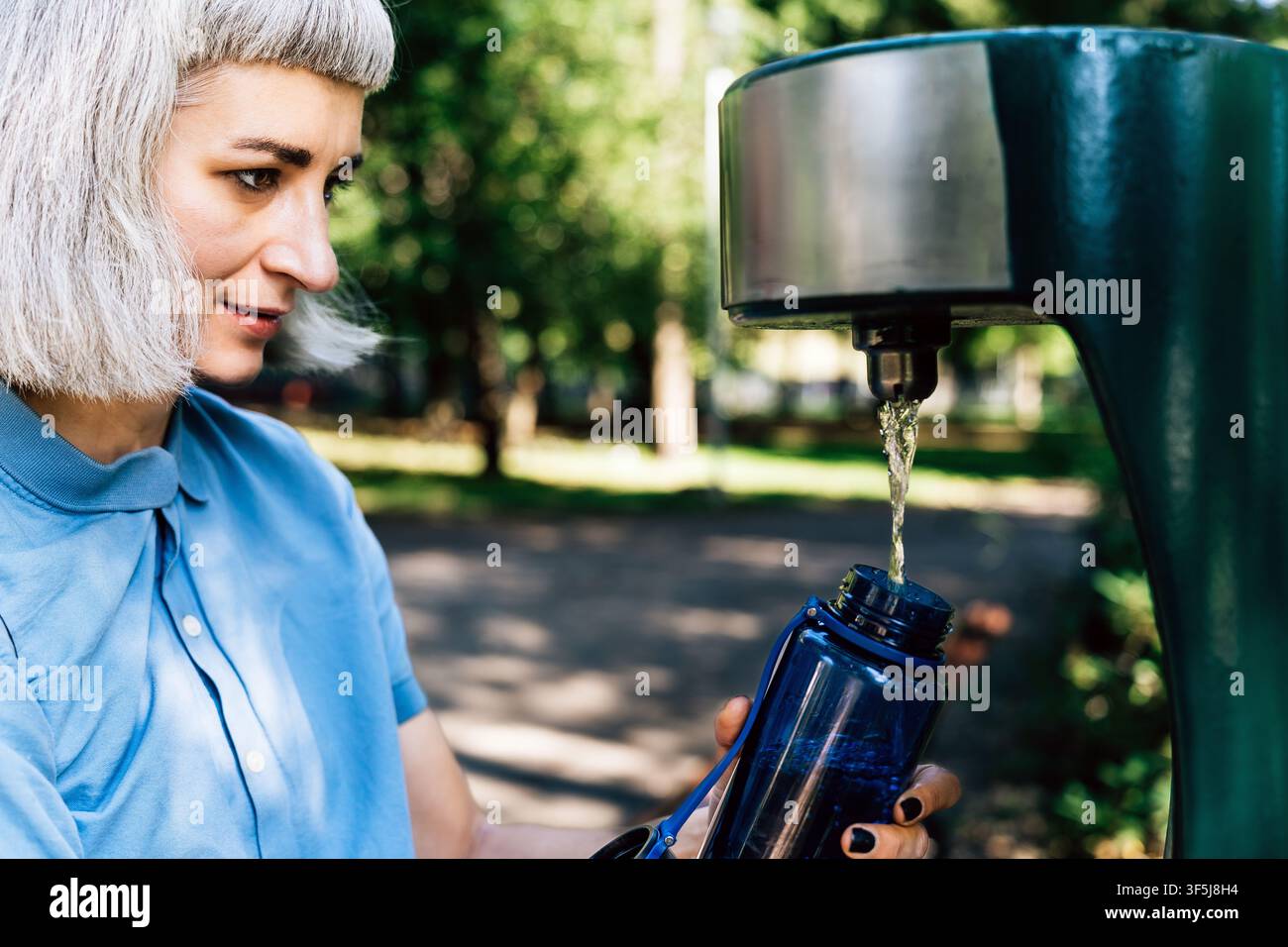 Gros plan d'une jeune femme aux cheveux gris courts remplissant une bouteille d'eau réutilisable à une fontaine publique dans un parc ensoleillé, promouvant l'hydratation un Banque D'Images