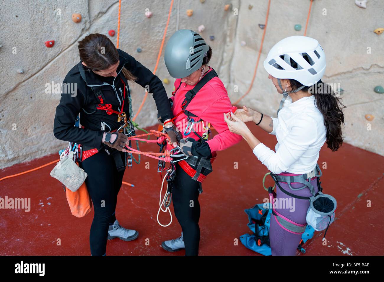 Trois femmes portant des casques et des harnais préparent des cordes et de l'équipement de sécurité lors d'un entraînement d'escalade et de spéléo en intérieur sur un mur de salle de sport. Banque D'Images