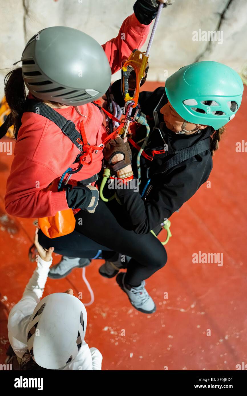 Trois femmes pratiquent des techniques de sauvetage vertical lors d'un entraînement à l'escalade en intérieur, utilisant des casques, des harnais, des cordes et des mousquetons à côté d'un cli artificiel Banque D'Images