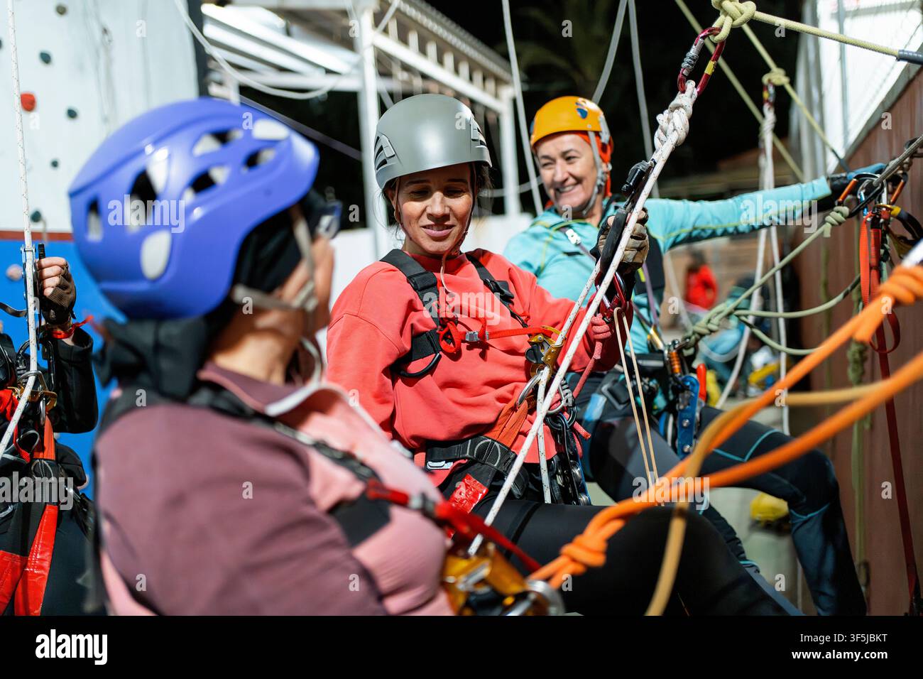Trois femmes pratiquent des techniques de spéléo vertical dans une salle de gym intérieure, portant des casques et des harnais tout en utilisant des cordes, des mousquetons et des équipements de sécurité pour Banque D'Images