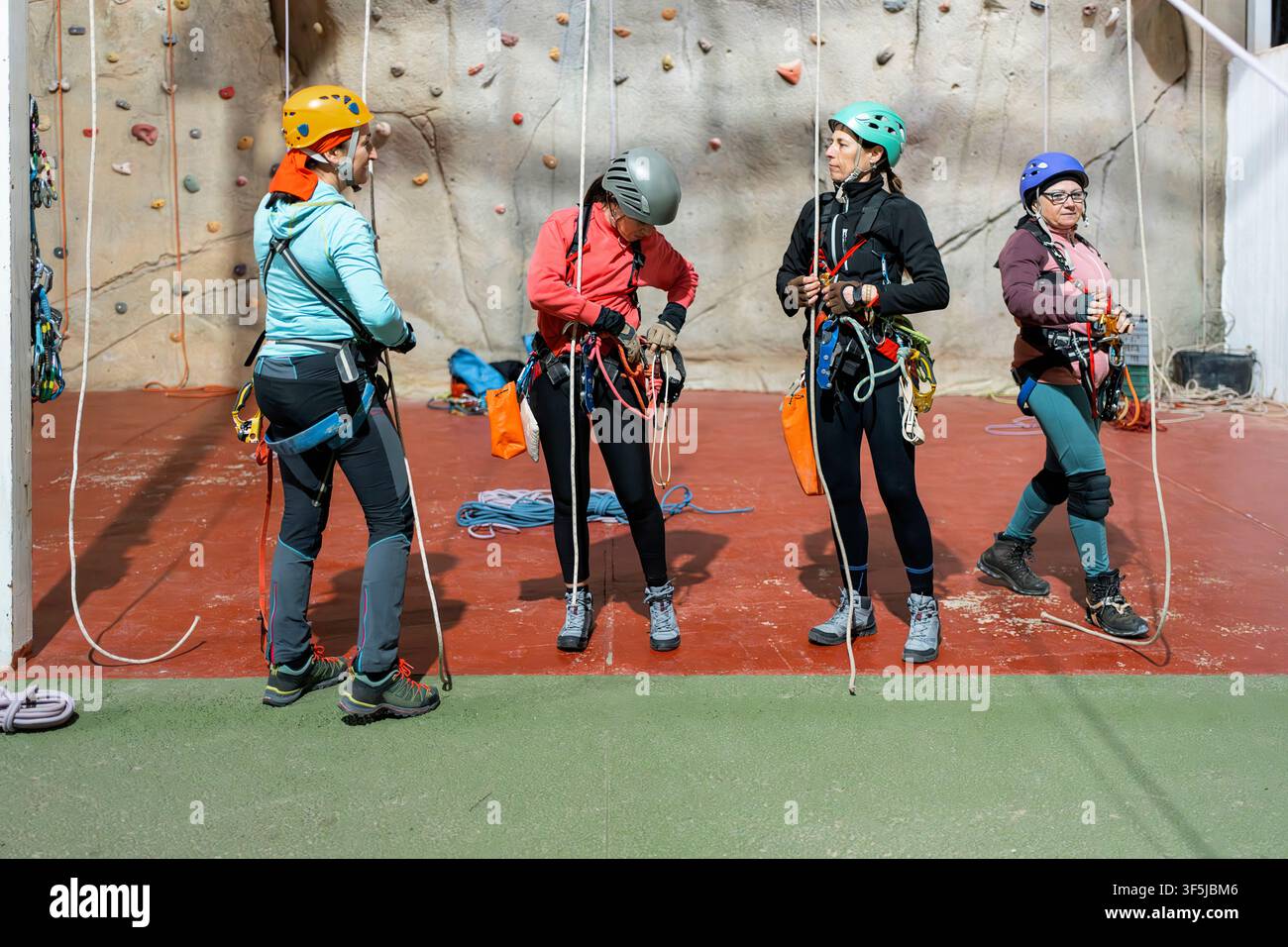 Quatre femmes casquées et équipées de harnais préparent des cordes et du matériel de sécurité lors d'un entraînement en escalade et spéléologie en intérieur sur un mur de salle de sport, pratiquant la manipulation verticale des cordes Banque D'Images
