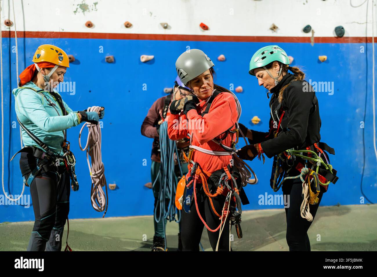 Trois femmes emballent des cordes et inspectent des harnais après une séance d'entraînement spéléologique en intérieur, portant des casques et des équipements de sécurité à côté d'un mur d'escalade coloré Banque D'Images