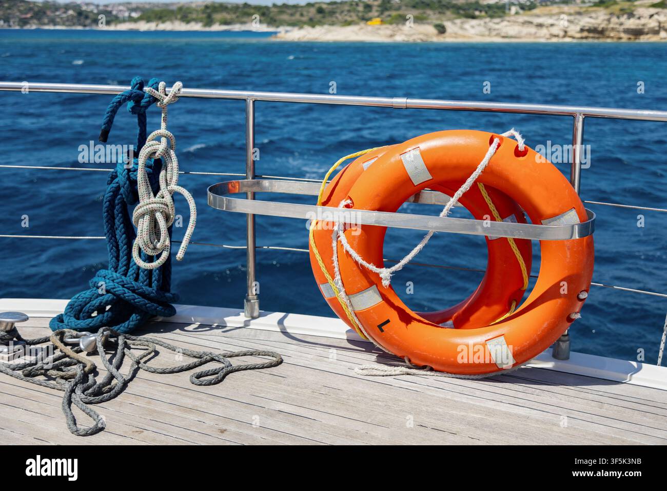 Bouée de sauvetage orange vif fixée à une rambarde en acier inoxydable sur le pont d'un yacht, avec une corde marine enroulée au premier plan et une mer bleue claire le long d'une roche Banque D'Images