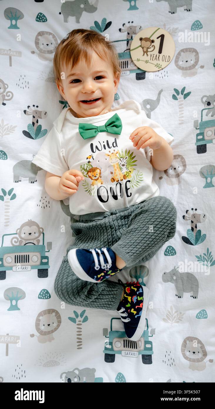 Garçon tout-petit souriant allongé sur une couverture sur le thème du safari, portant une chemise blanche "Wild One" avec un nœud papillon vert et tenant une carte de jalons pour le premier anniversaire pour la première bi Banque D'Images