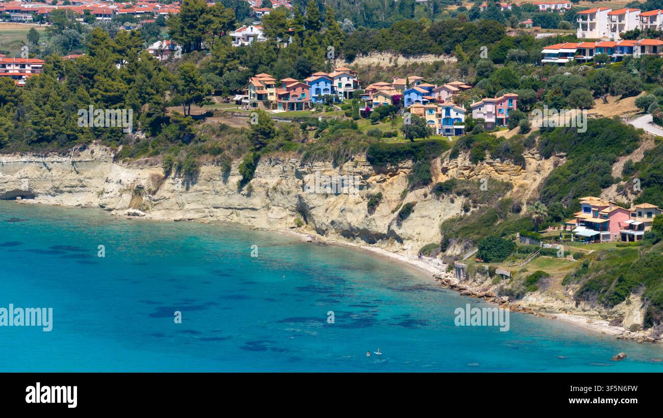 Skala Fourkas, Grèce, 11 juillet 2026. La mer turquoise rencontre des falaises rocheuses claires en dessous d'appartements et maisons résidentielles colorés entourés d'une végétation dense Banque D'Images