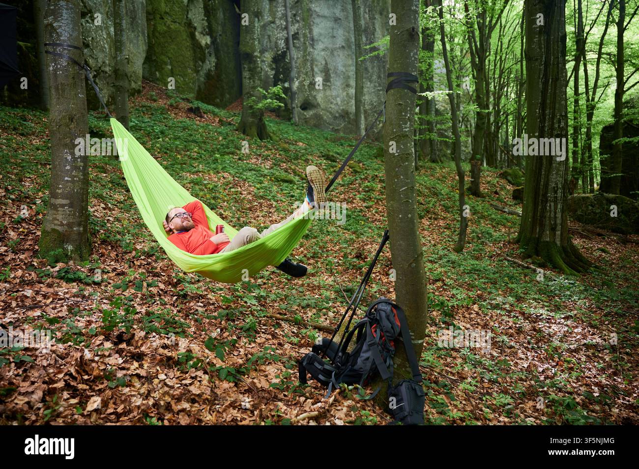 Personne se relaxant dans un hamac vert vif attaché entre deux arbres dans la forêt luxuriante et verte près des rochers. Homme touriste tient à boire, profitant d'un moment paisible entouré par la nature. Sac à dos et équipement de repos à proximité. Banque D'Images